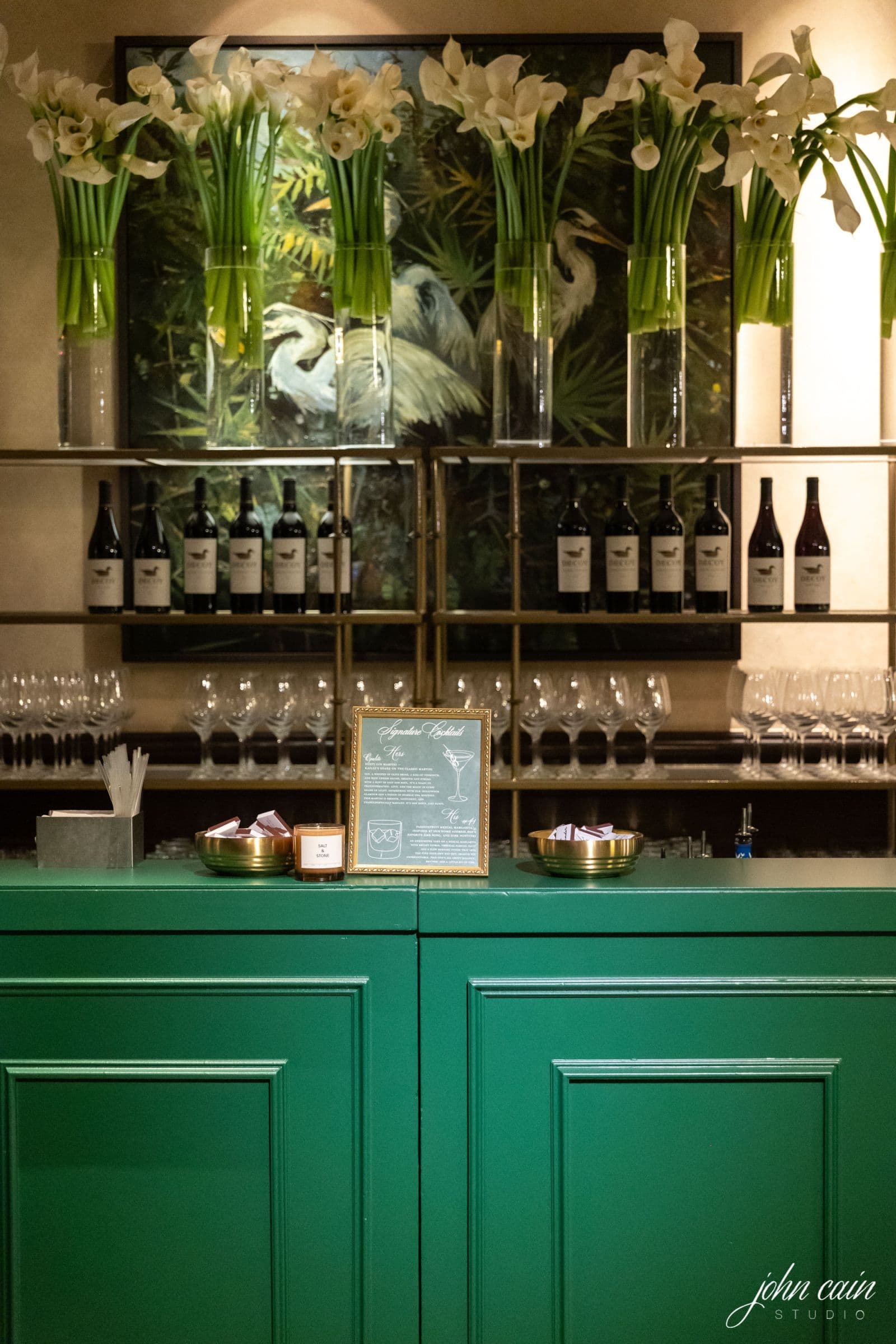A green bar with white flowers, wine bottles, and a signature cocktail menu.