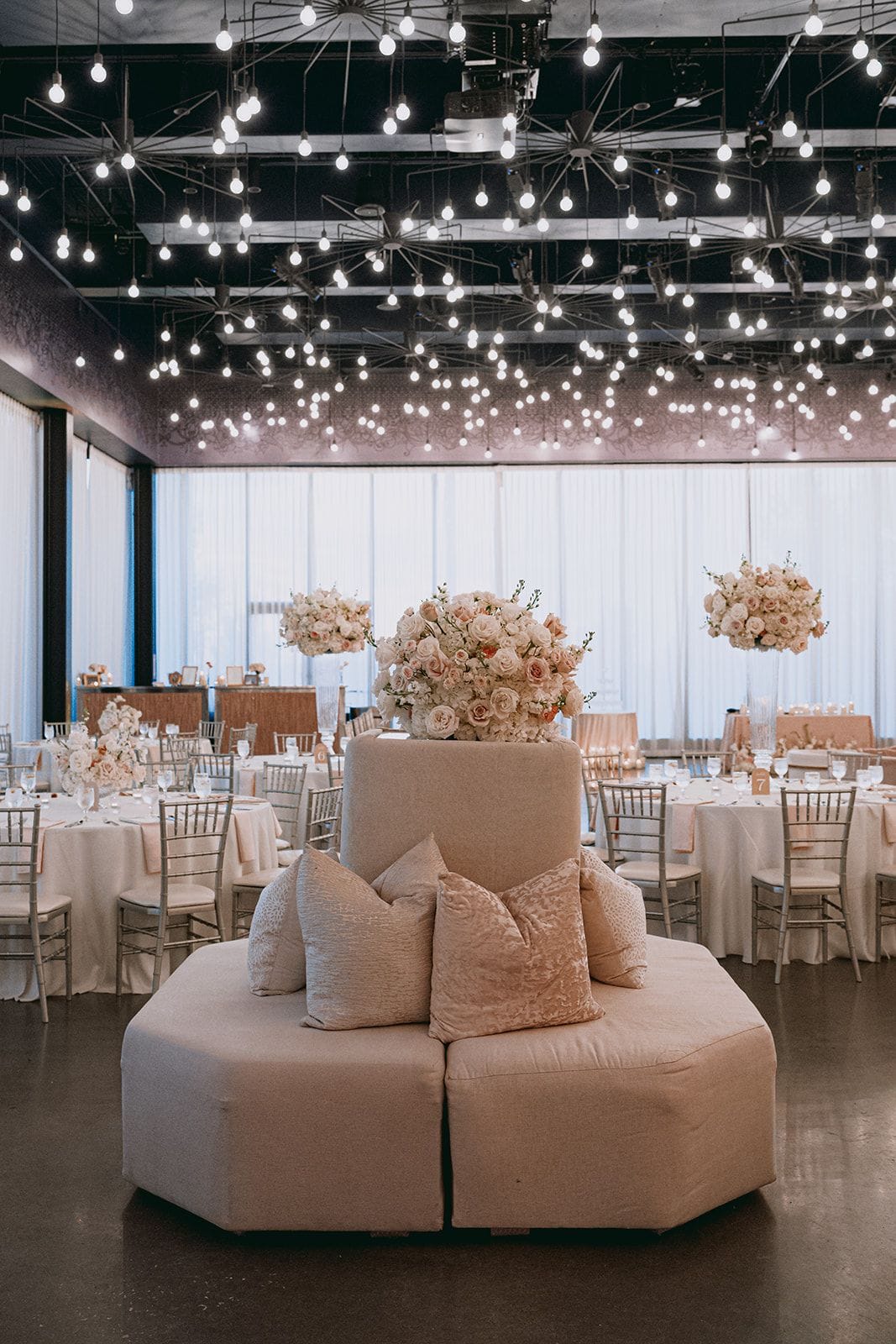 A wedding reception hall with round tables, floral centerpieces, and a lounge area.